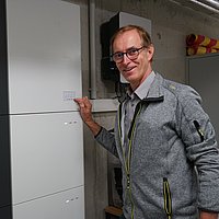 Stefan Römmele zeigt mit der Hand auf den Wärmespeicher im Technik-Raum des Gemeindezentrums.